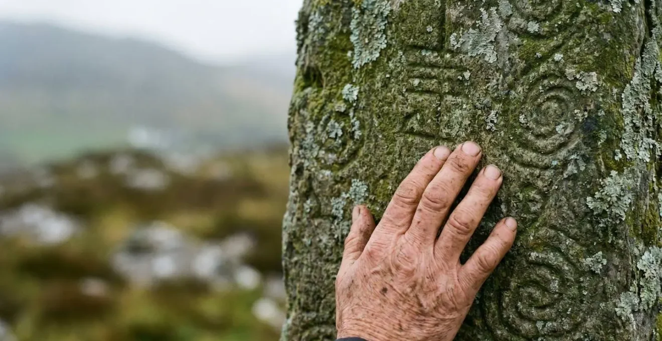 A weathered hand gently touching ancient Celtic stone carvings with Ogham inscriptions in soft natural light